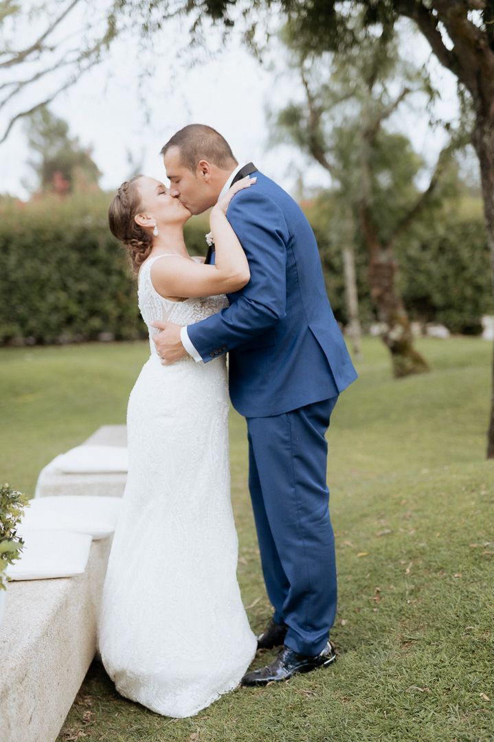 A bride in a white lace gown and groom in navy suit share a romantic kiss on a grassy lawn at an outdoor wedding venue.