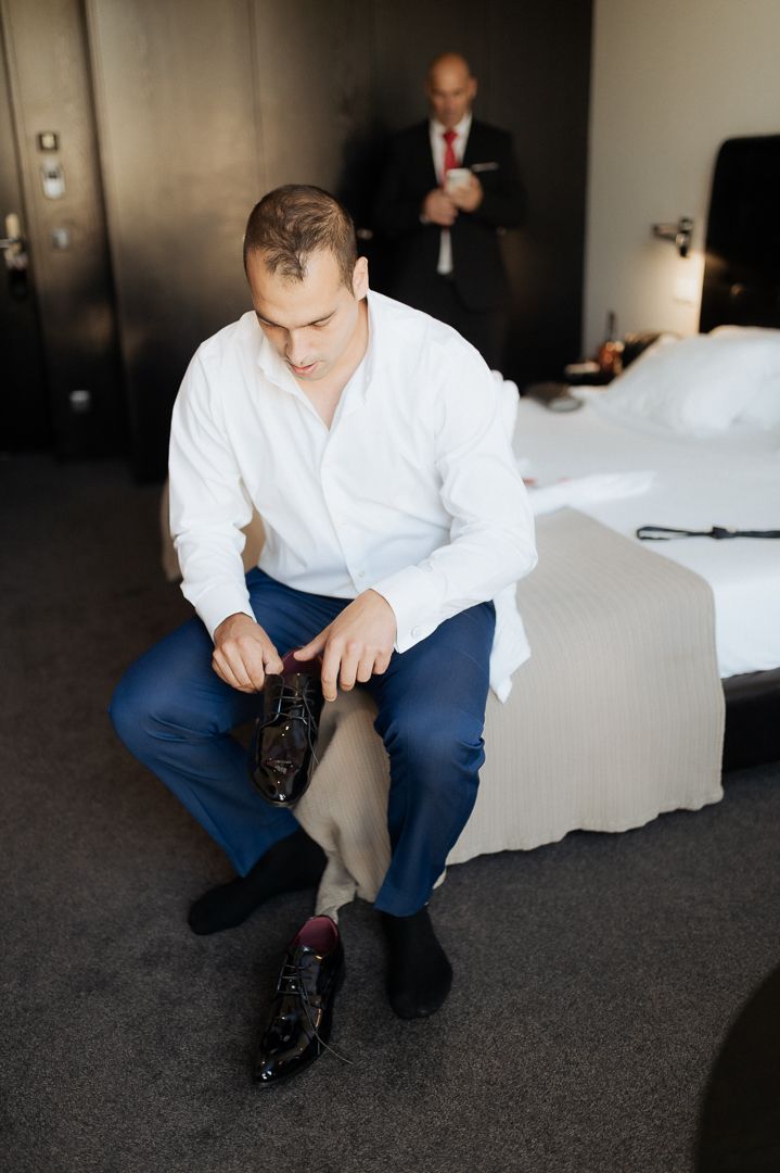 A groom in white shirt and navy pants sits on a bed tying his formal black dress shoes.