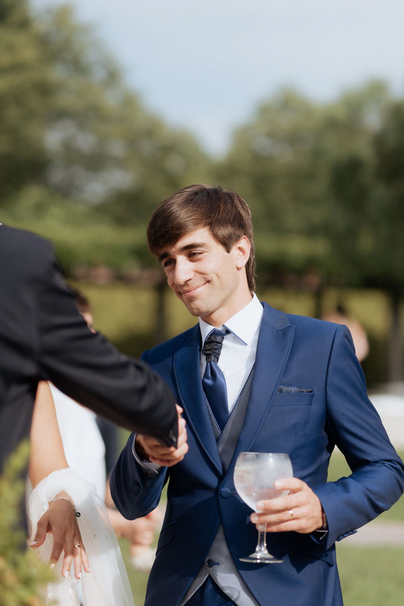 Guest in a navy blue suit holds a wine glass while socializing at an outdoor wedding reception.