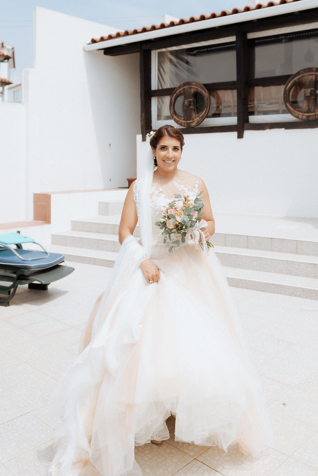 Someone wearing a beautiful white wedding dress stands on outdoor steps holding a delicate bouquet of pastel flowers.