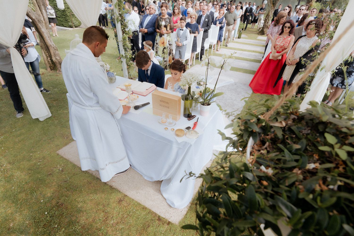 An outdoor wedding ceremony with a priest at a table conducting the service while guests watch from the lawn.
