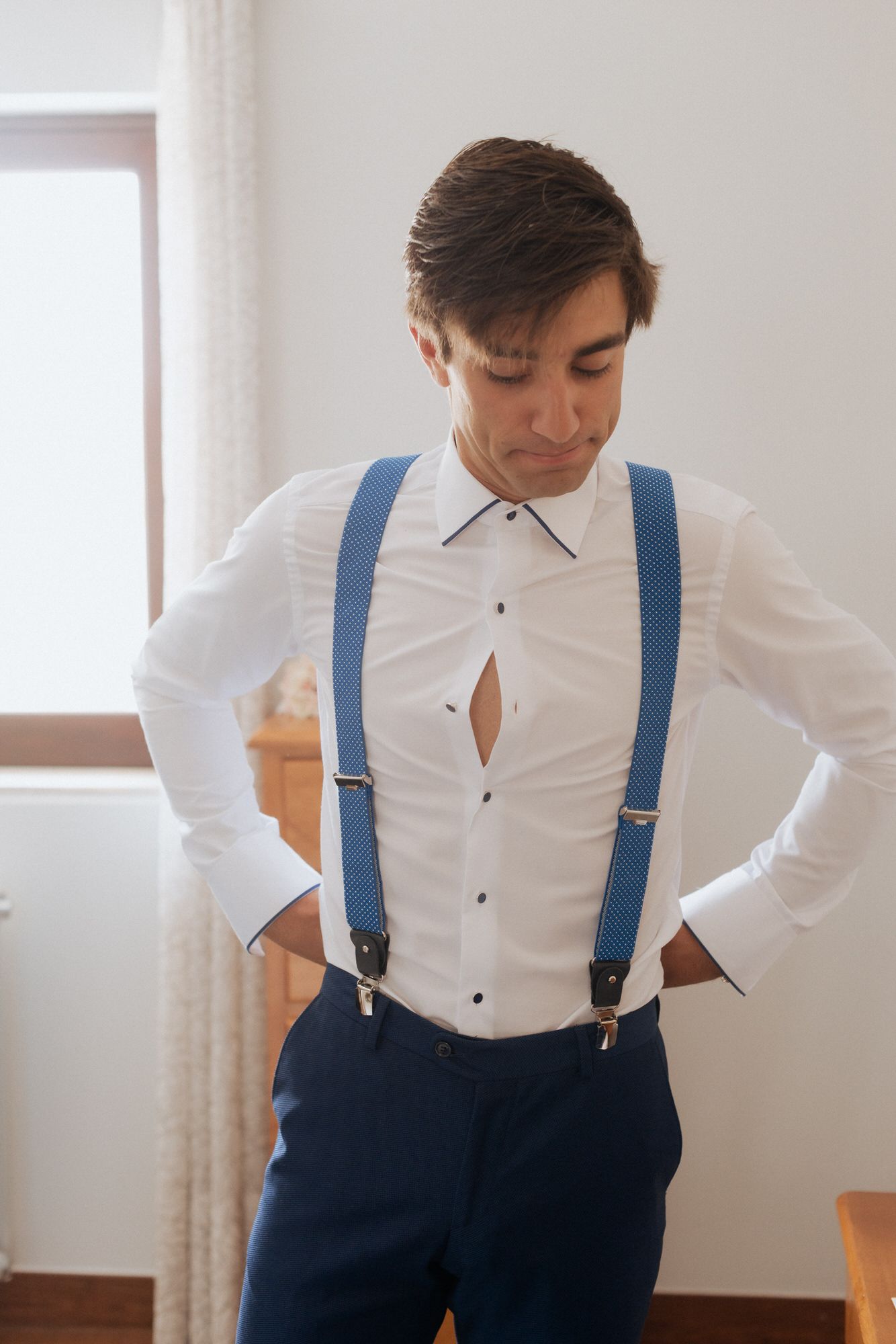 Close up view of a groom wearing a crisp white dress shirt with blue suspenders and navy pants.