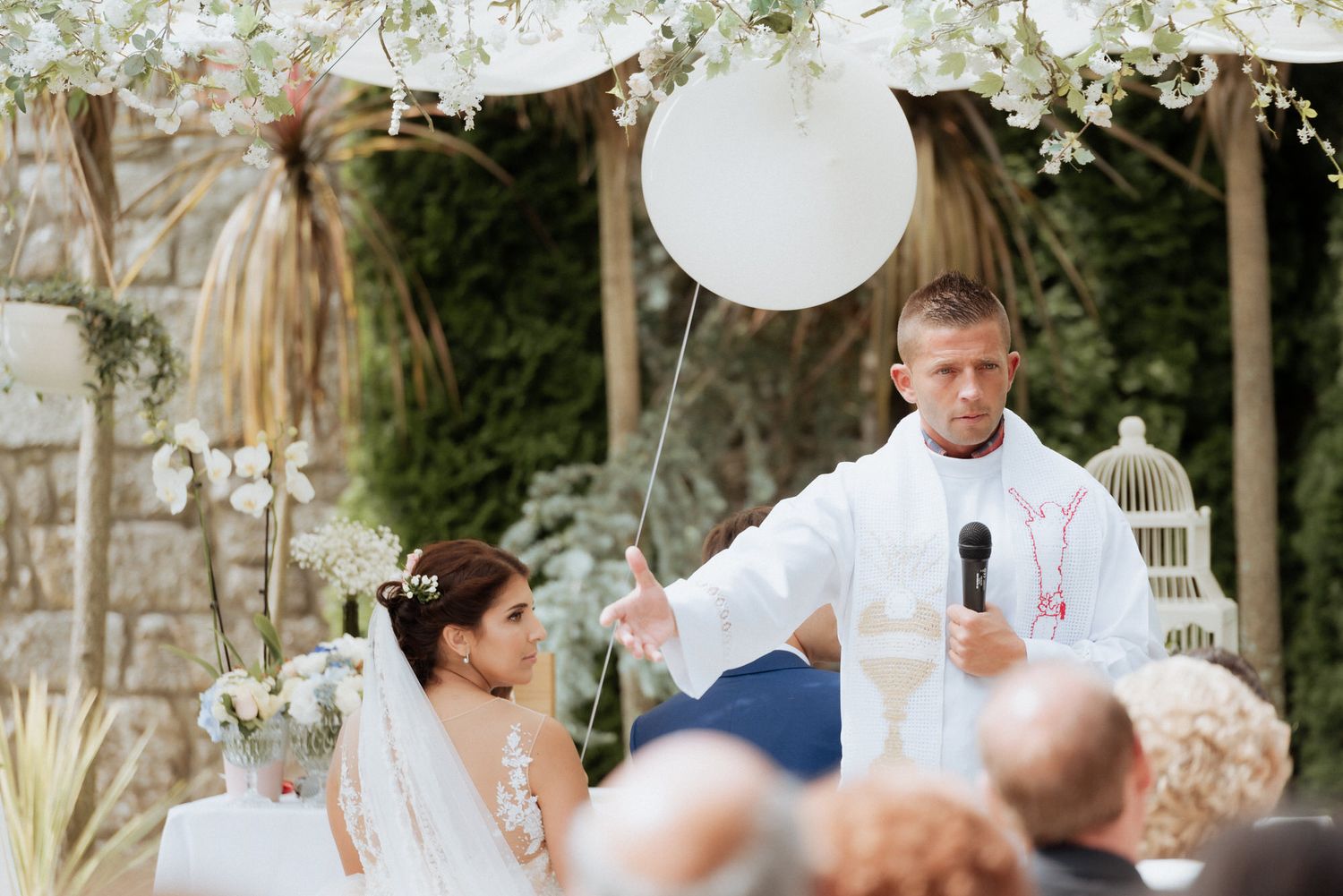 Wedding reception speech under an outdoor tent decorated with white lanterns and floral arrangements during a daytime celebration.