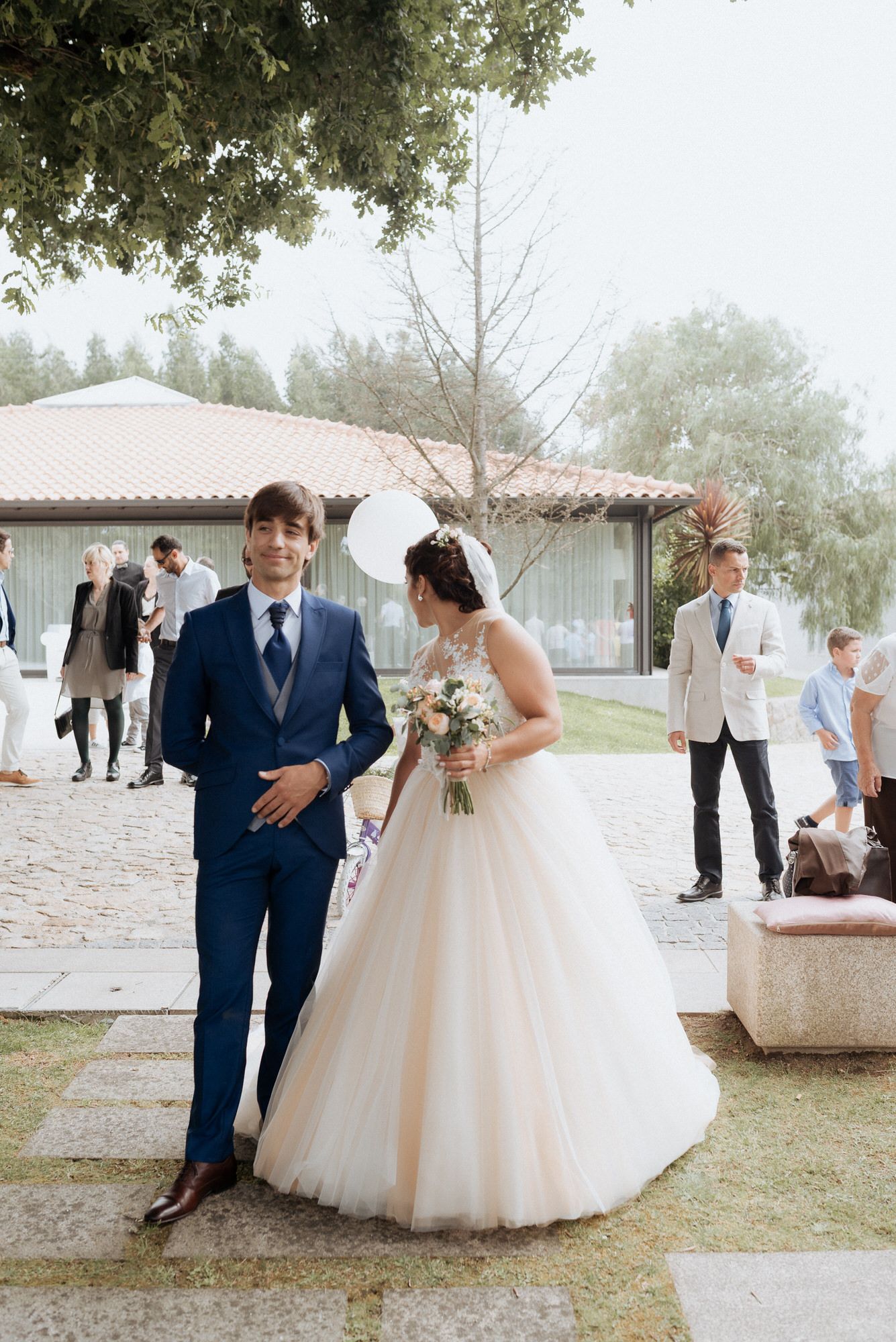 Wedding celebration featuring a bride in a flowing white ballgown and groom in navy suit at an outdoor venue.