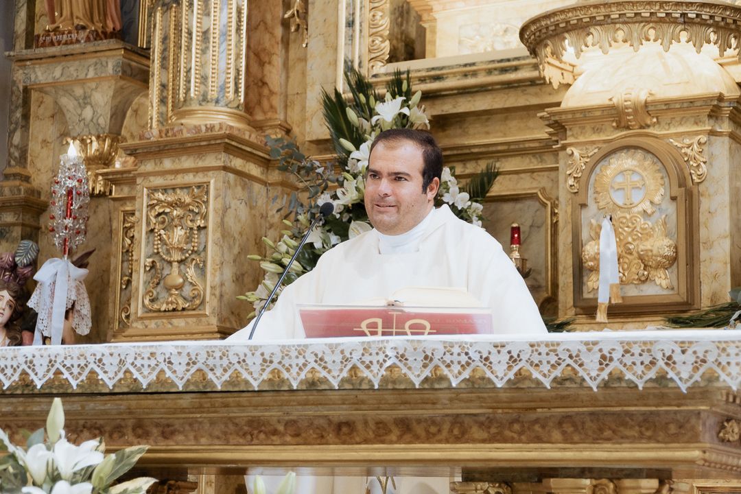 Priest in white robes stands at ornate golden altar during religious ceremony.