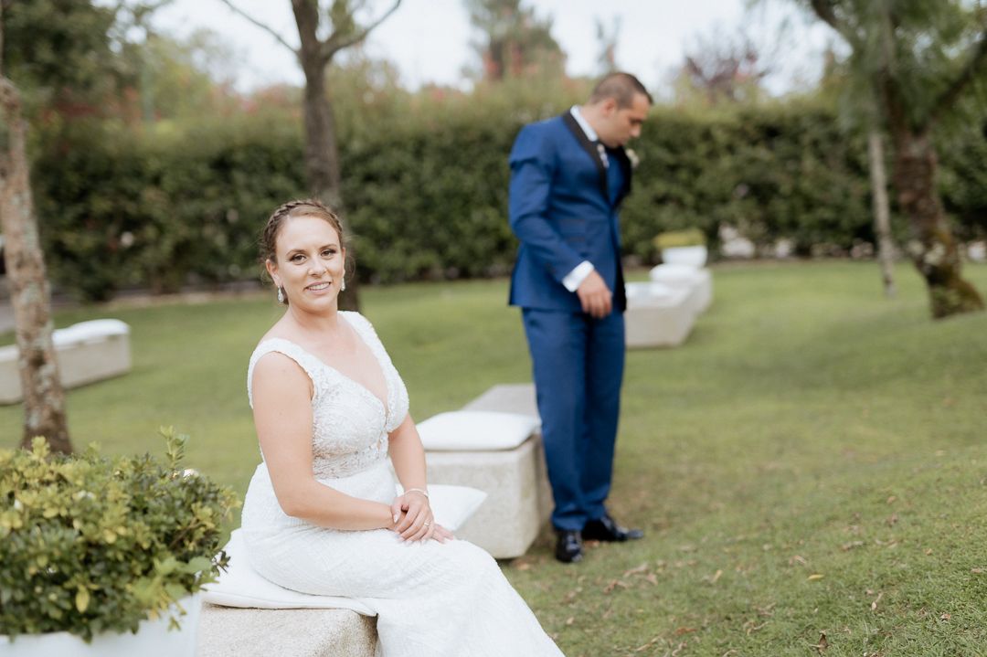 A bride in a white dress sits on a bench in a garden while a groom in a blue suit stands in the background.