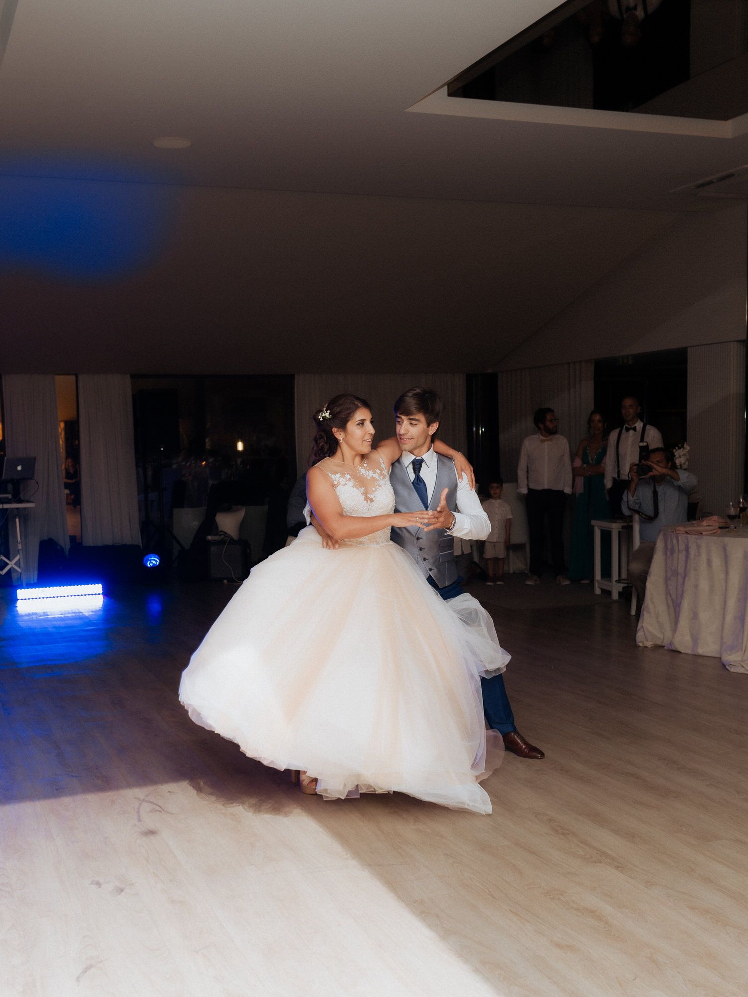 A couple shares their first dance at their wedding reception, with the bride in a flowing white ballgown on a light wooden floor.