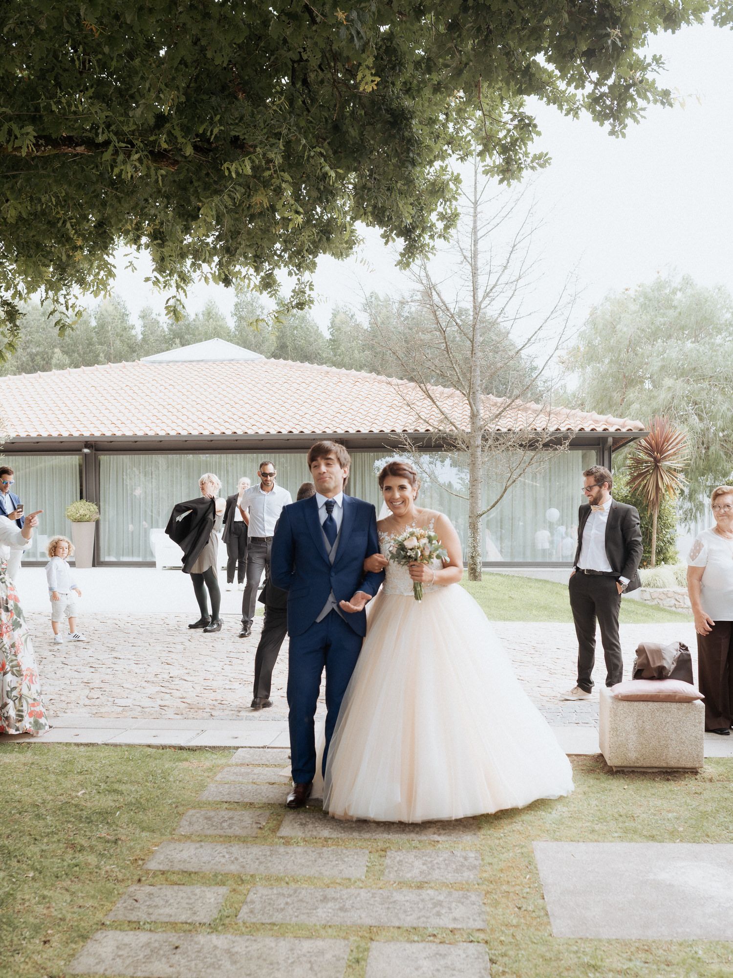 Wedding celebration at an outdoor venue with white stucco building, stone pathway and lush trees in the background.