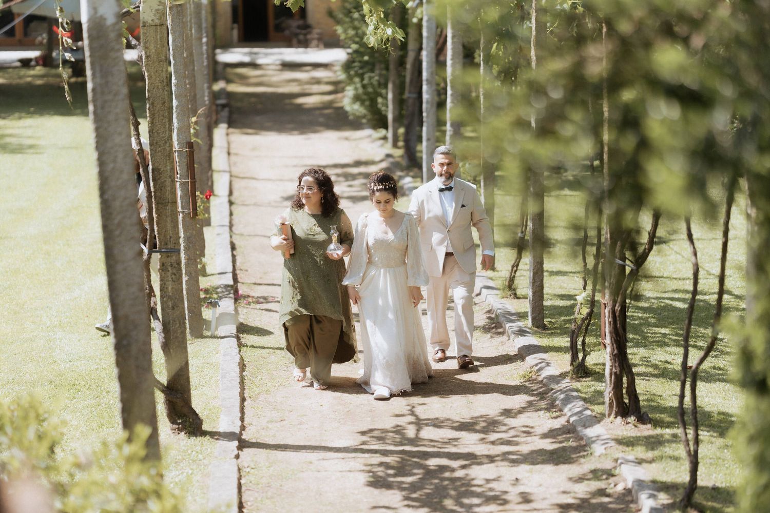 A wedding party walks down a sunlit garden path lined with hanging decorations and greenery.