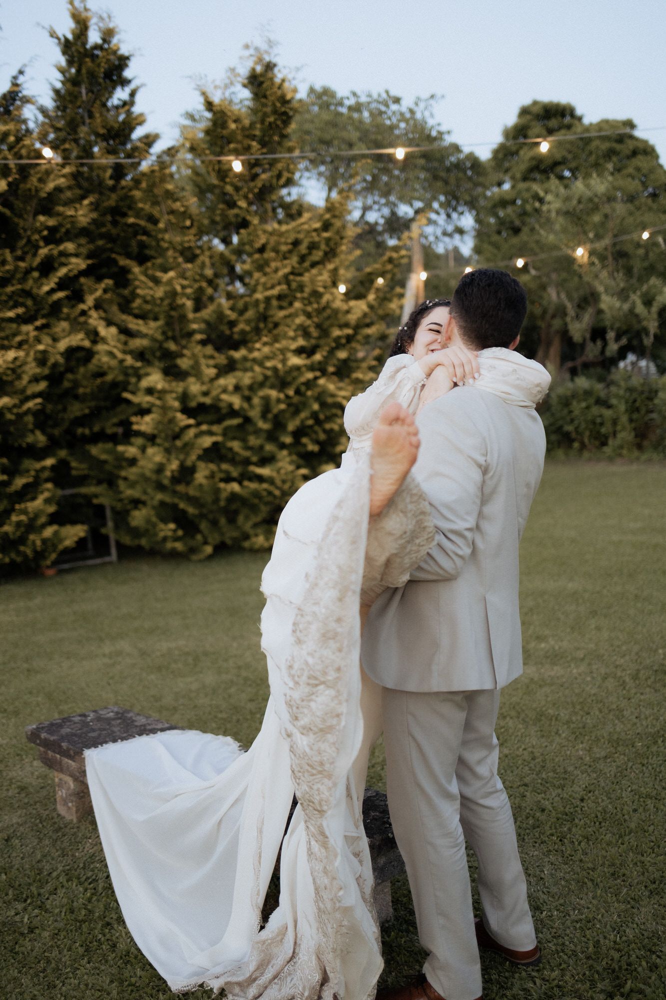 A couple embraces outdoors at sunset, with the bride wearing a lace wedding gown and the groom in a light colored suit.