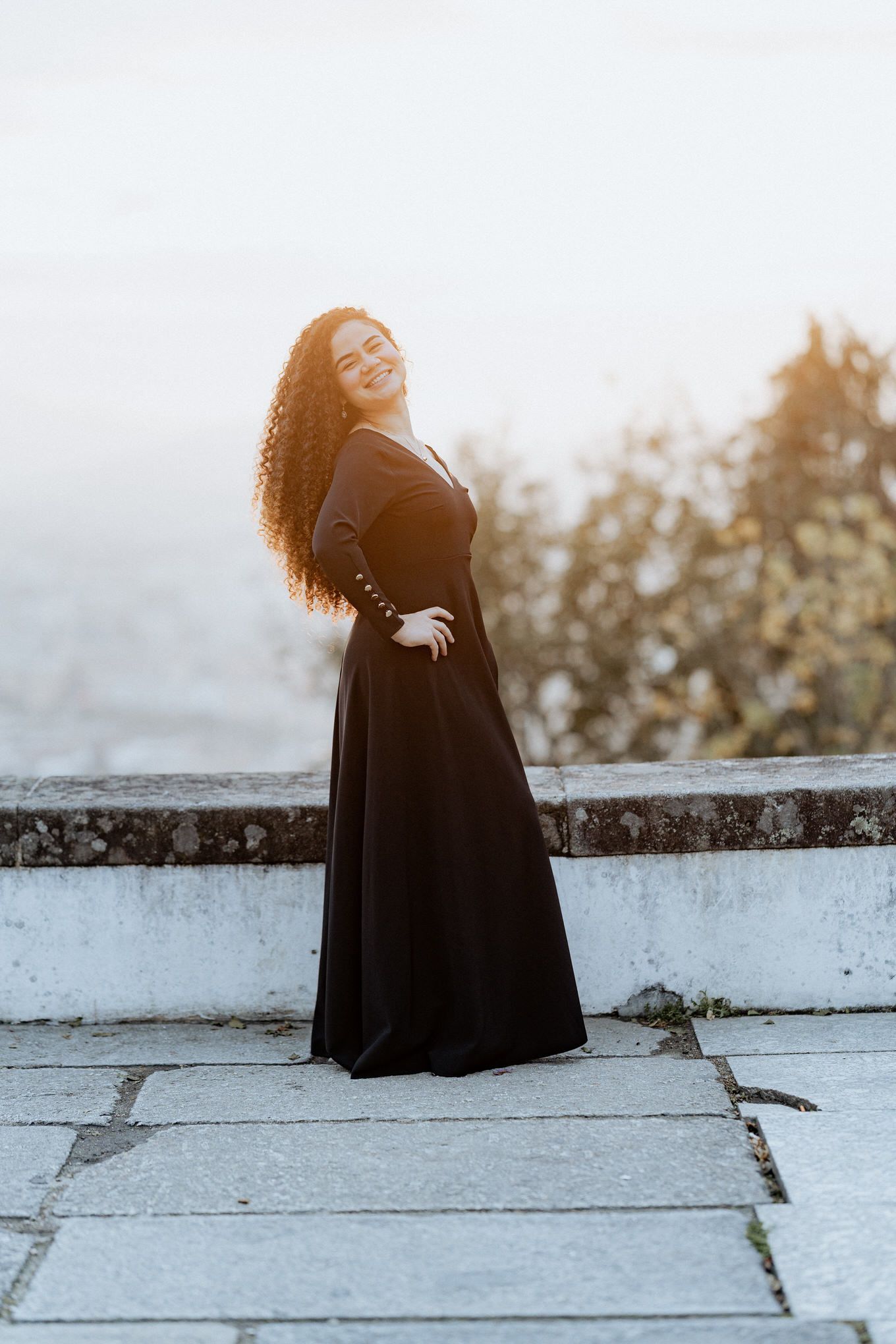 Uma mulher em um vestido preto esvoaçante posa elegantemente contra uma parede de pedra durante a hora dourada.