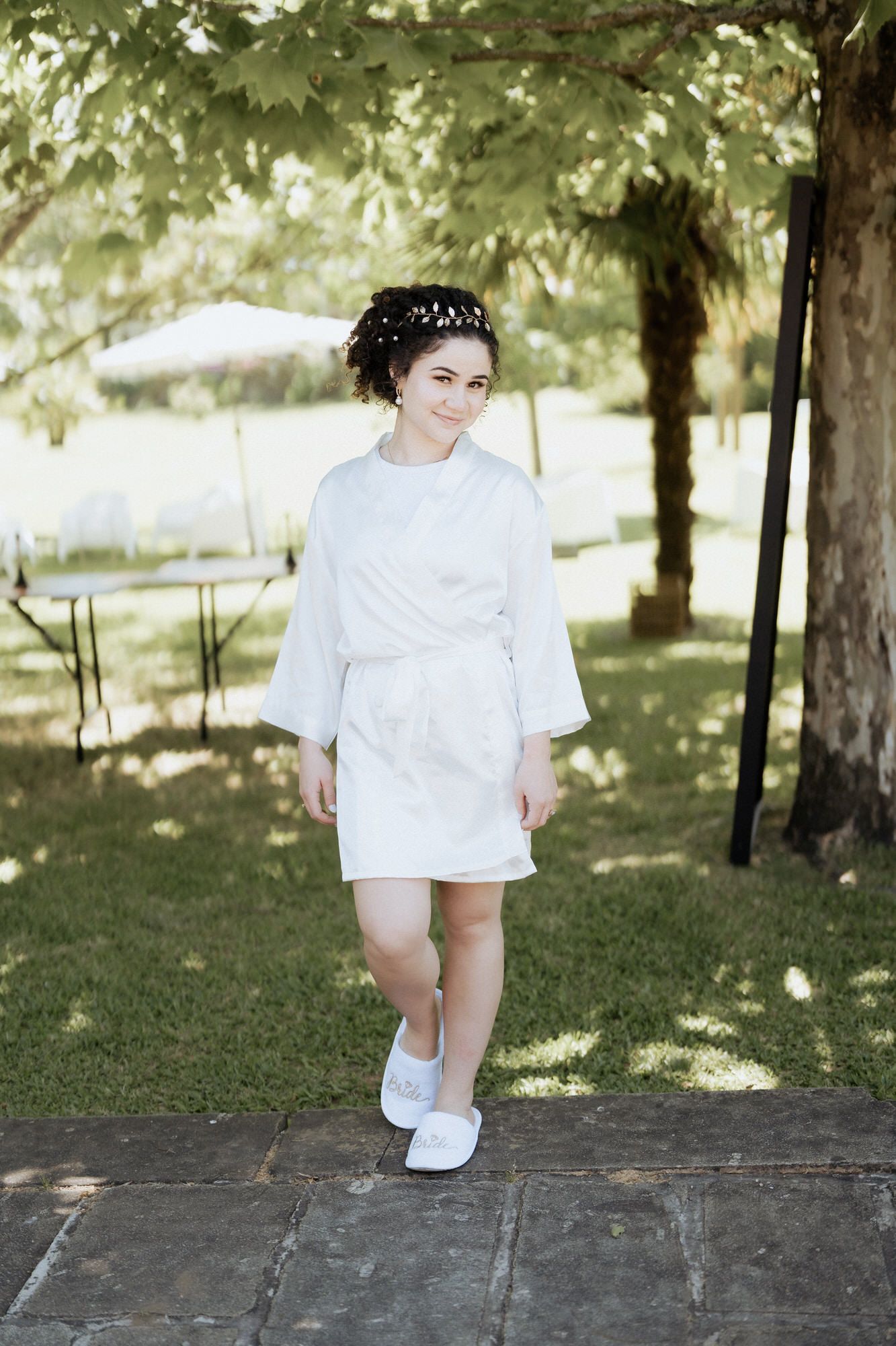 Someone in a white spa robe and slippers stands on stone pavers under a shady tree in a peaceful garden setting.