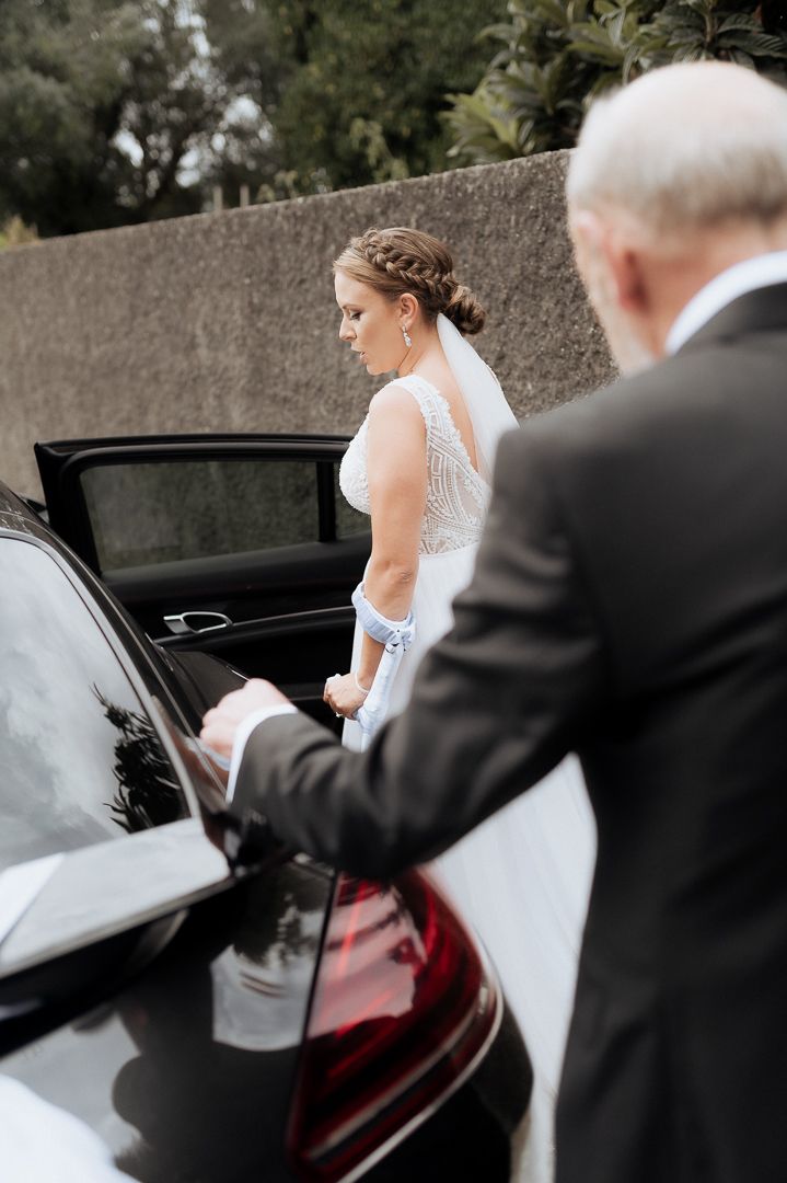 Elegant wedding scene with bride in lace dress entering luxury black car while accompanied by guest.