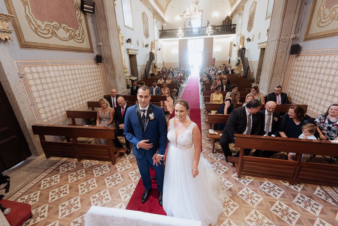 A couple stands at the front of an ornate church with guests seated in wooden pews behind them during a wedding ceremony.