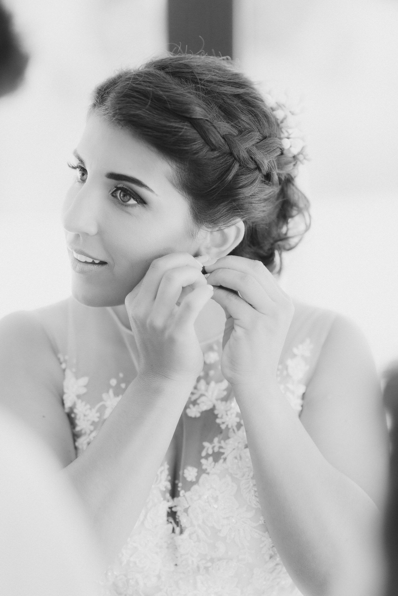 Black and white photo of a bride putting on earrings, wearing a delicate lace wedding dress with a braided updo hairstyle.