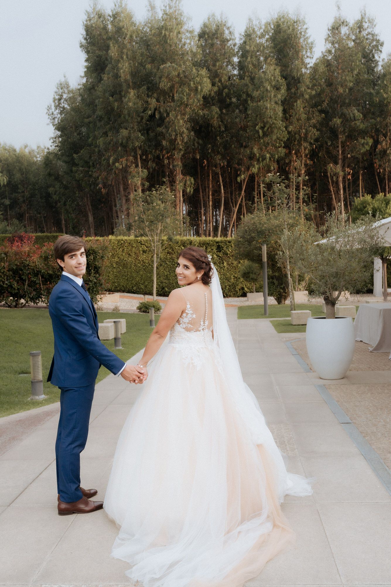 Wedding couple shares a romantic first dance moment outdoors in a garden setting with olive trees and modern planters.