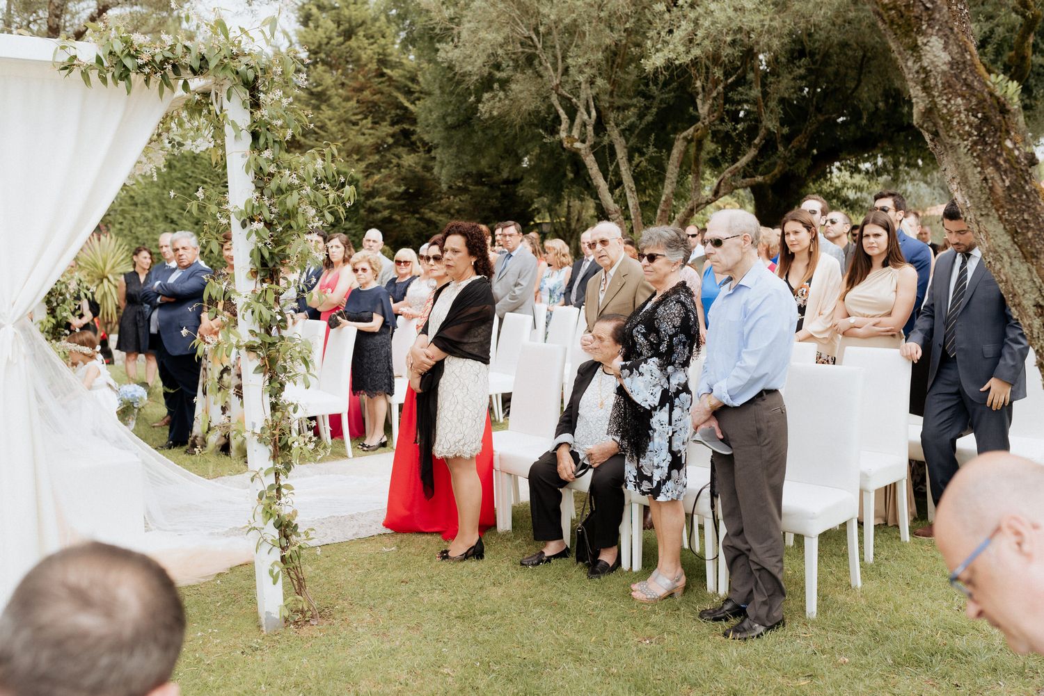 Wedding guests standing in an outdoor garden ceremony space with white chairs and floral arrangements under trees.