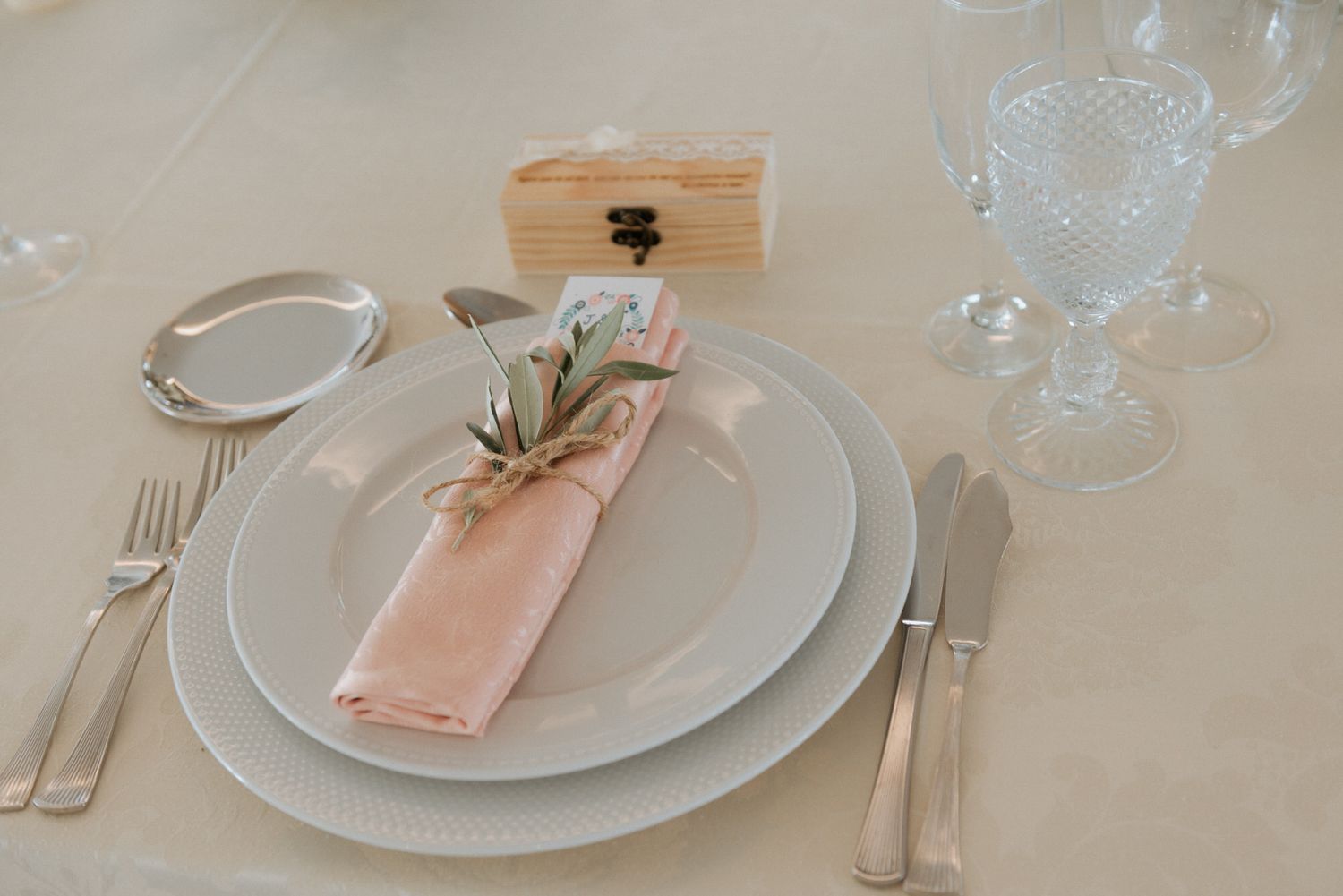 An elegant table setting with a peach napkin, rosemary sprig and crystal glassware on a cream tablecloth.