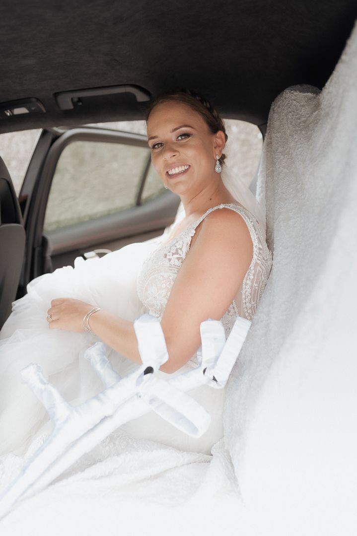 A smiling bride wearing a white lace wedding dress sits in the backseat of a car on her special day.