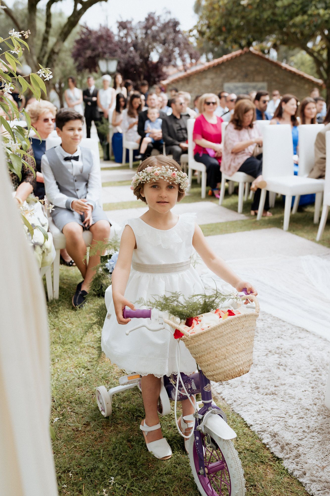 Child in a white dress rides a decorated bike with a basket during a wedding ceremony procession on a garden path.