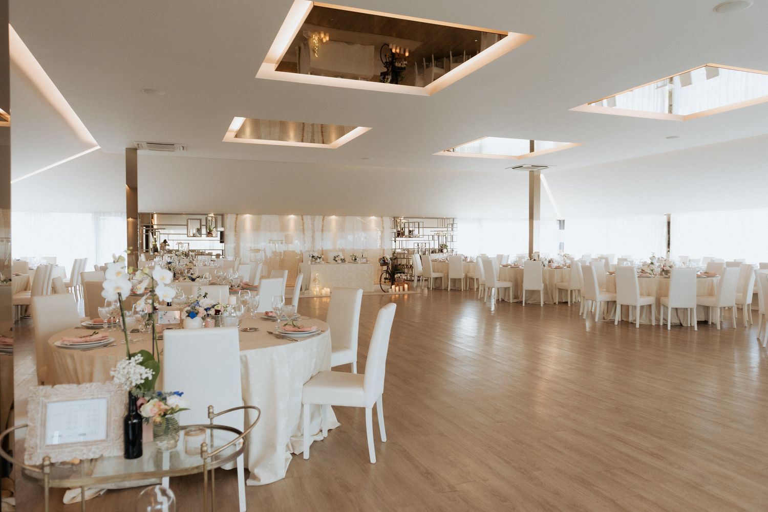 Elegant wedding reception venue with white tables, chairs, and modern lighting fixtures in a bright open space.