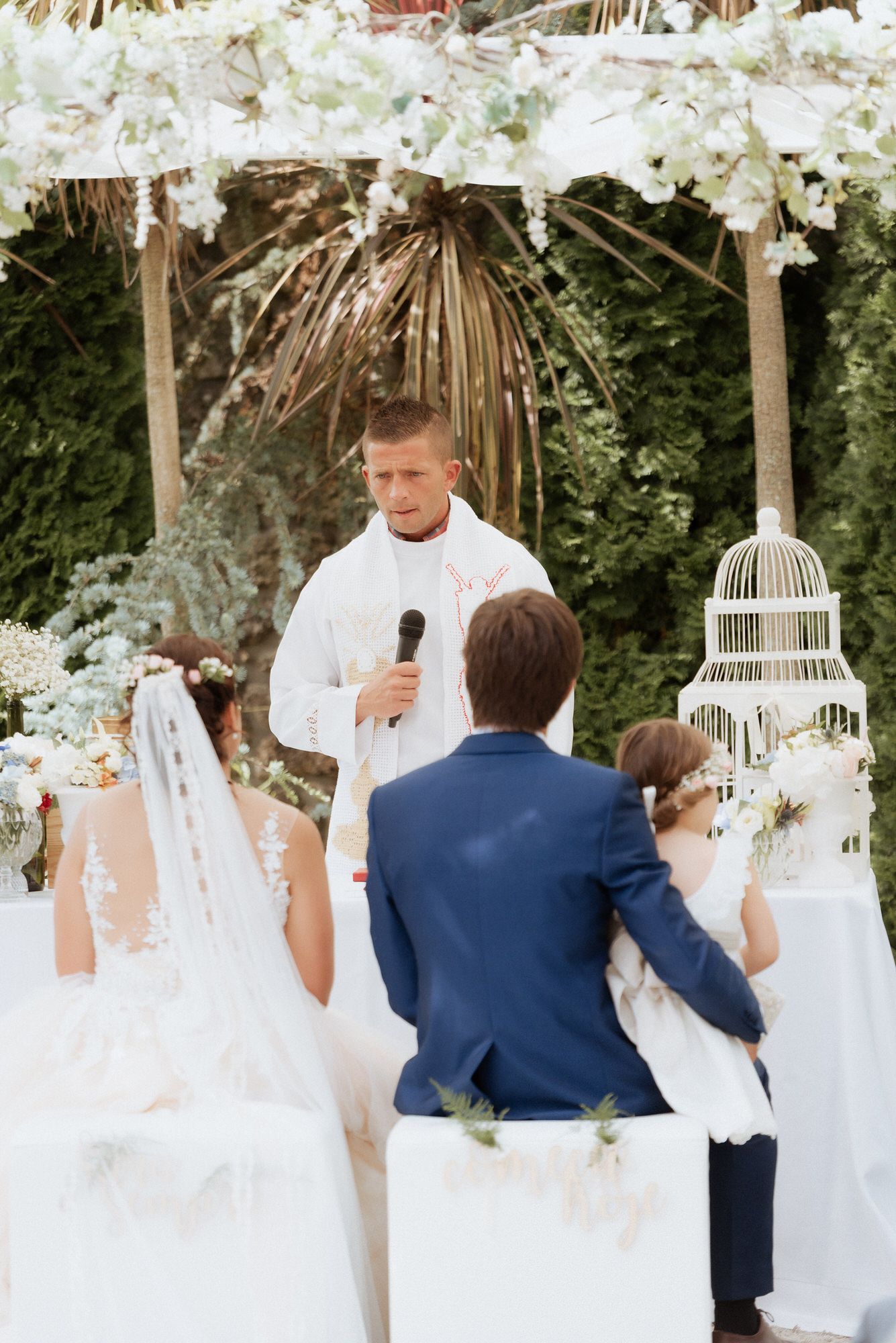 Wedding ceremony in a garden setting with an officiant speaking to the couple under a decorated arch with white flowers.
