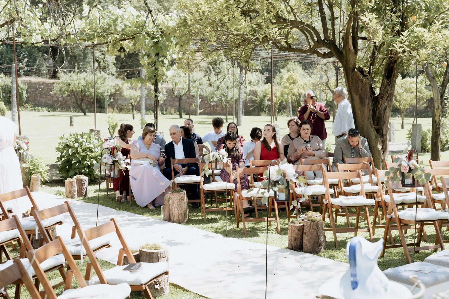 Wedding guests mingle at an outdoor garden reception with rustic wooden chairs and elegant table settings under shady trees.