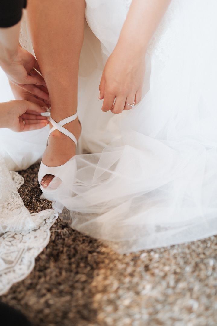 Delicate white ballet-style wedding shoes being fastened on gravel ground with tulle dress flowing around.