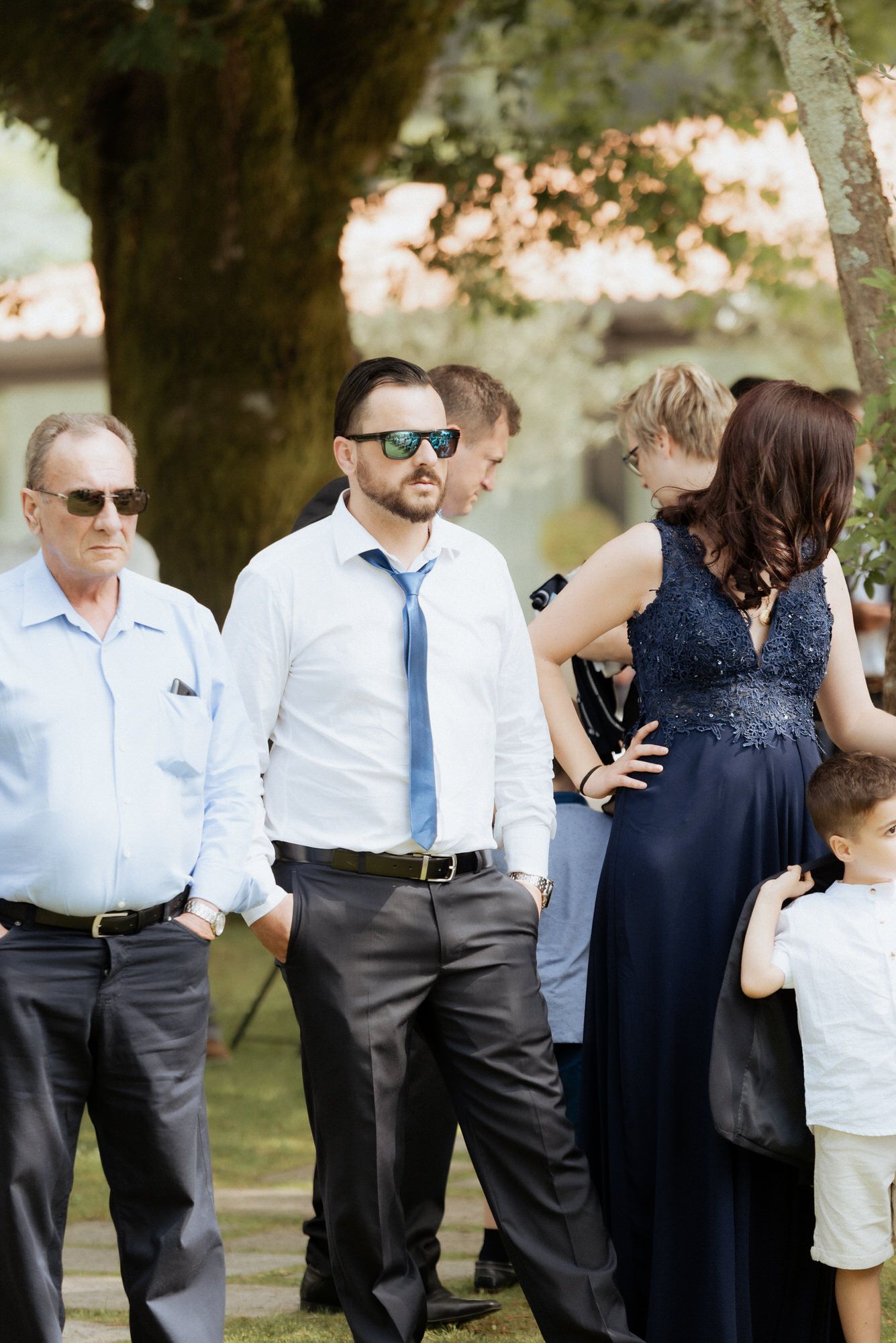 Guests dressed in formal attire gather outdoors under trees at an elegant event on a sunny day.