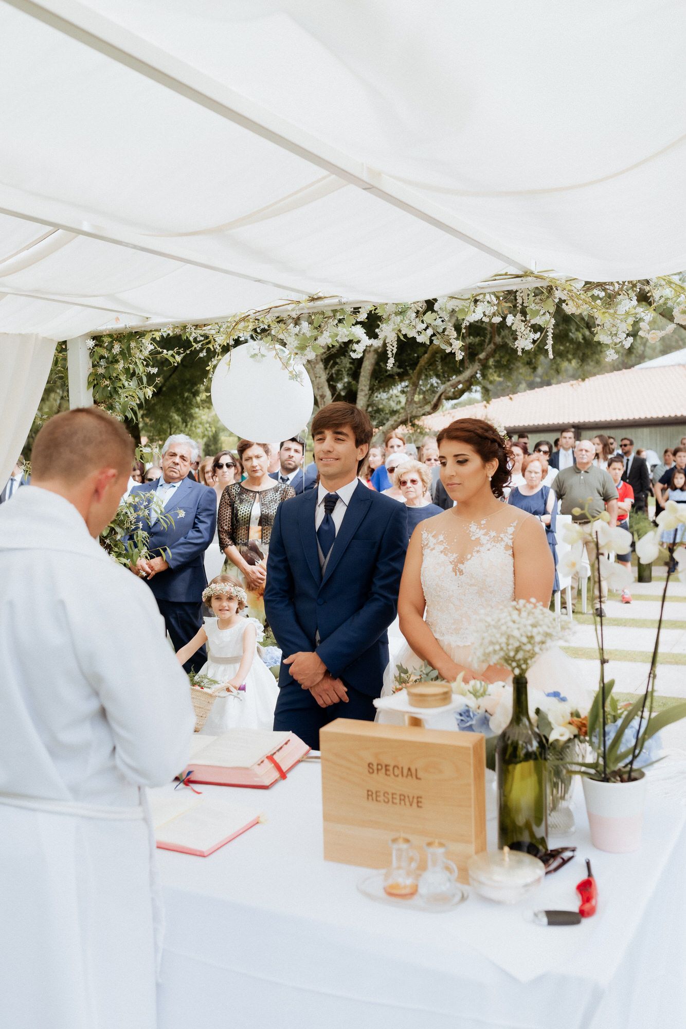 A couple stands at an elegant outdoor wedding ceremony under a white tent decorated with greenery and flowers.