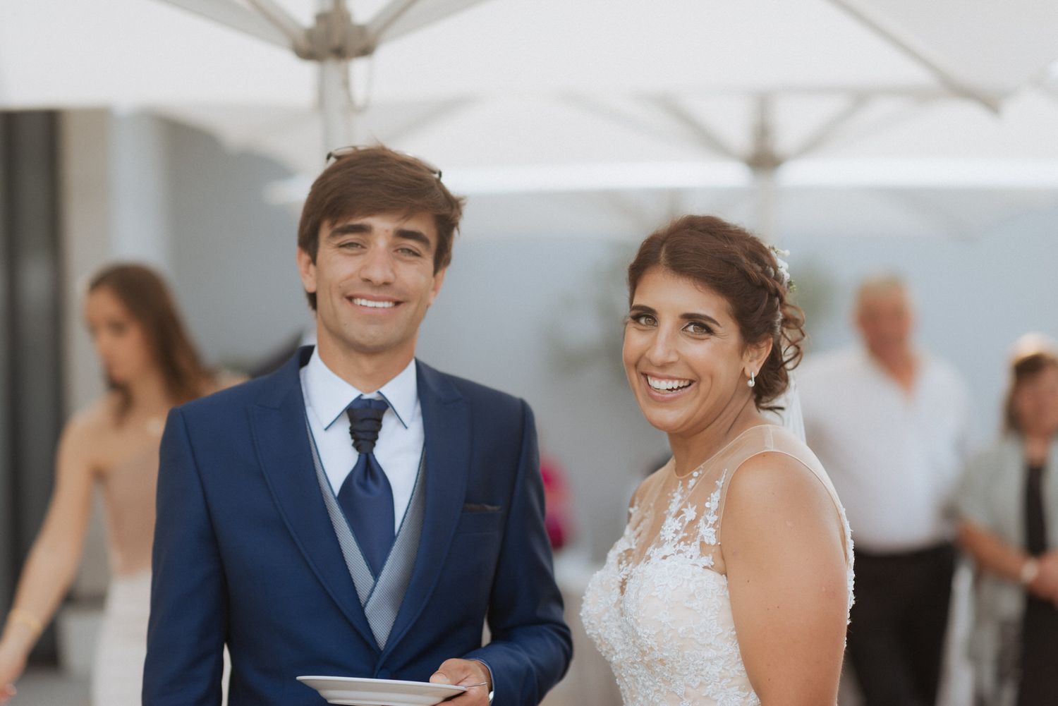 A smiling couple at an outdoor wedding celebration stand together under white umbrellas on their special day.
