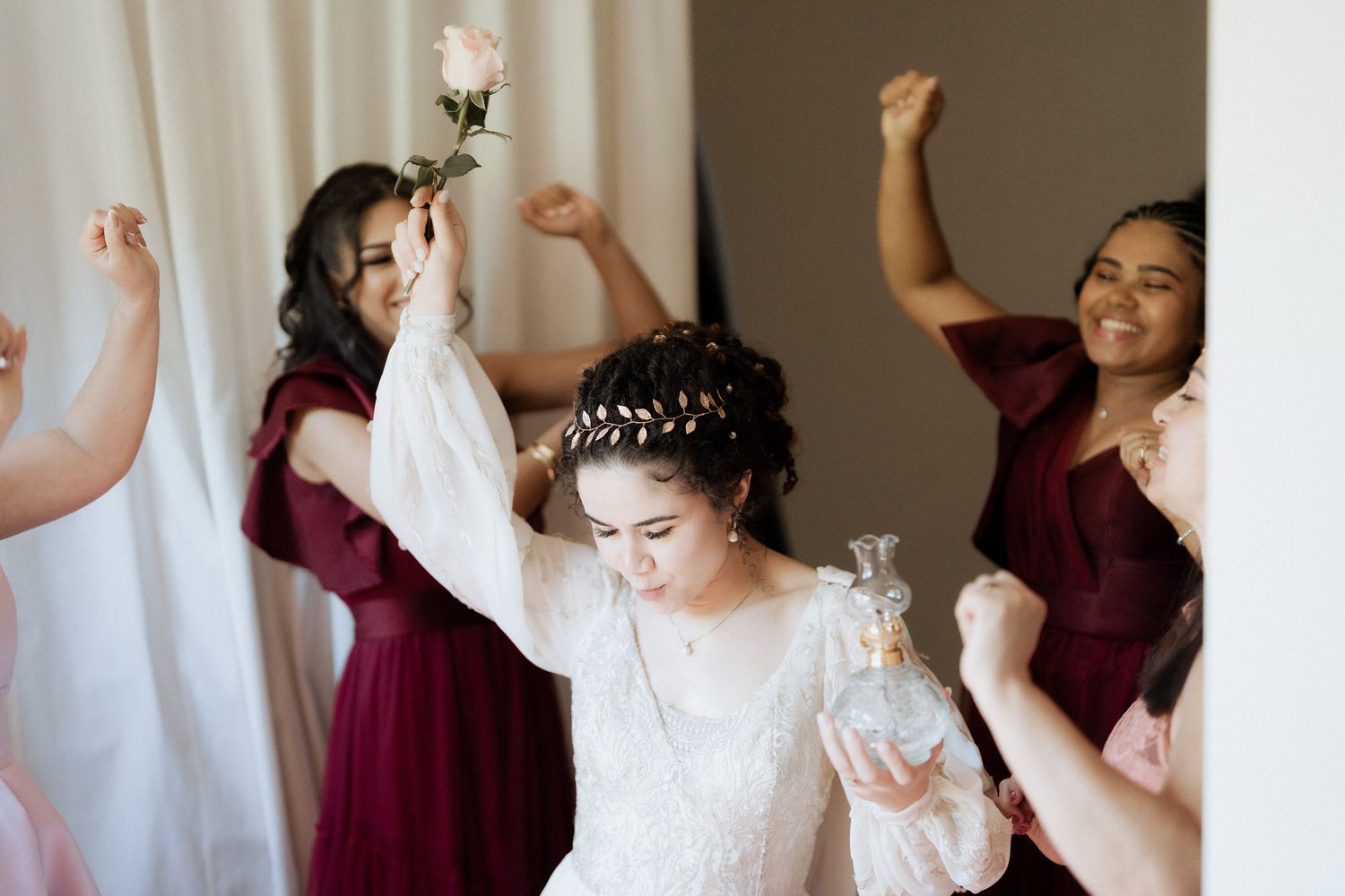 A group of bridesmaids in burgundy dresses celebrate joyfully with the bride in white dress and tiara.