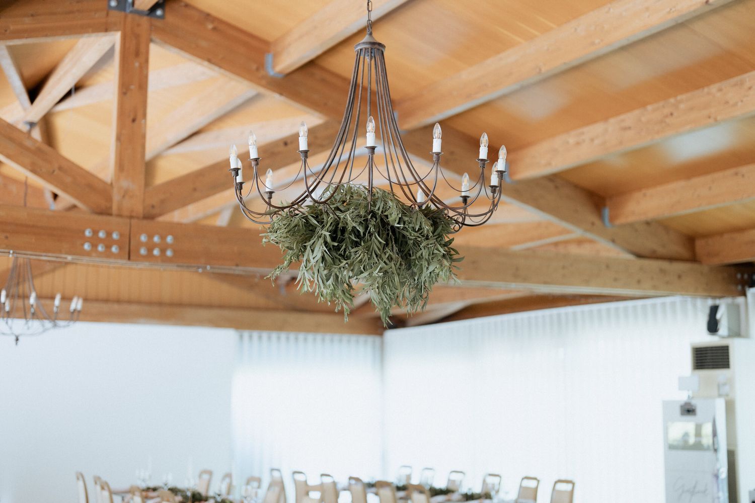 A rustic chandelier adorned with greenery hangs from wooden beams in an event venue space with white walls.