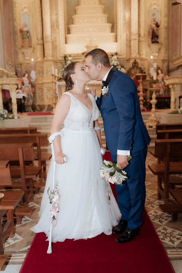A couple shares their first kiss during a wedding ceremony inside an ornate Catholic church with wooden pews and red carpet.