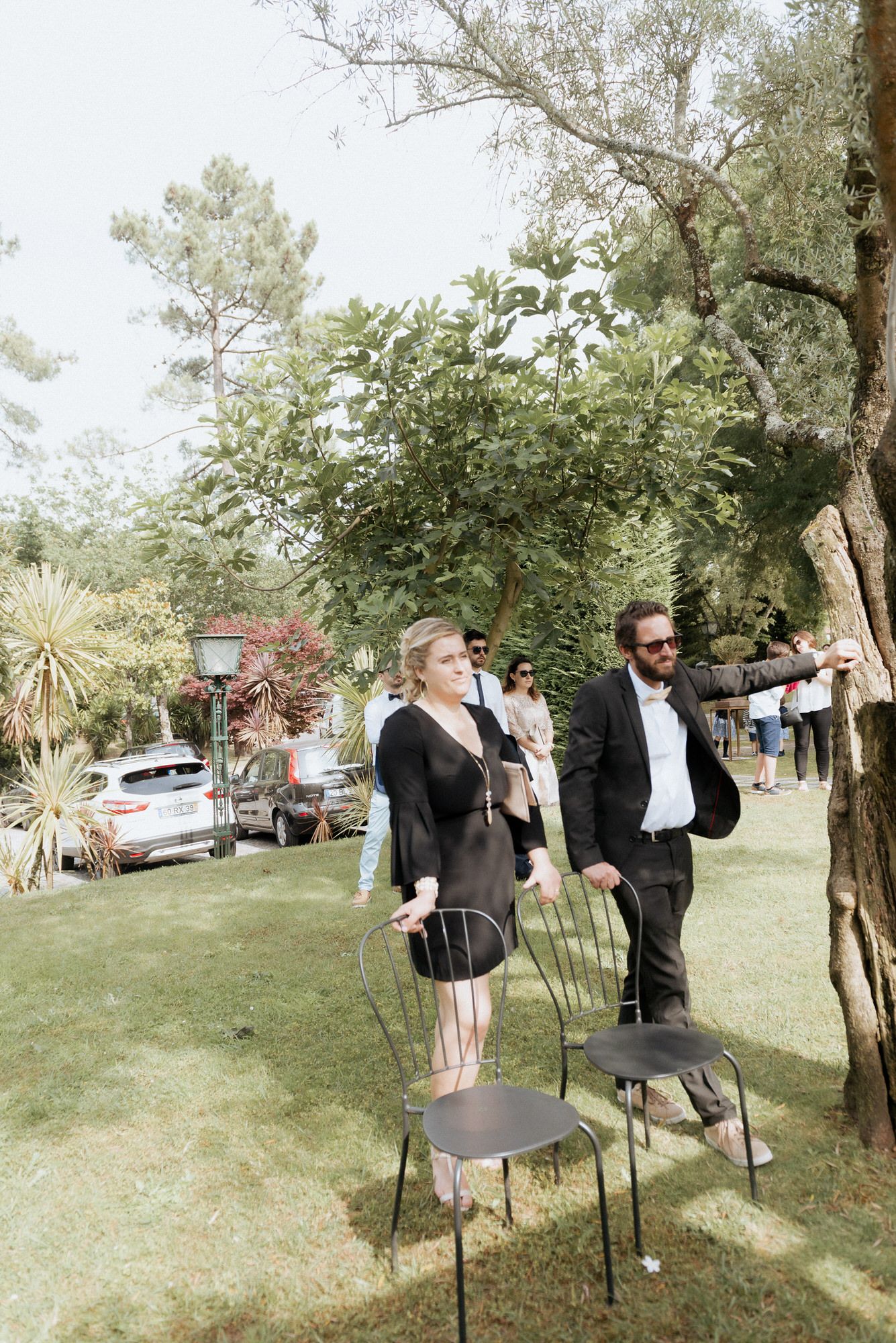 Two guests stand on chairs at an outdoor garden party surrounded by lush greenery and trees.