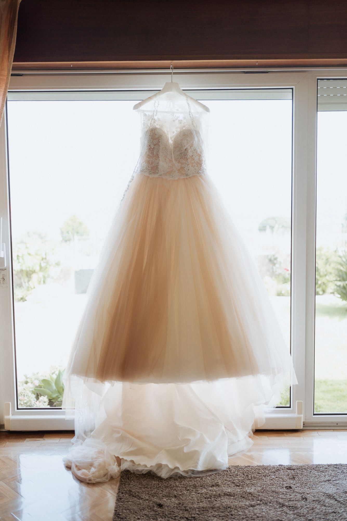 An elegant white wedding dress with lace bodice and tulle skirt hangs gracefully in front of a bright window.