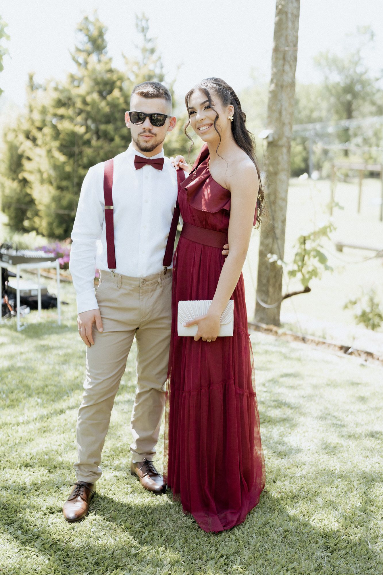 A couple poses on grass at an outdoor event, wearing a burgundy gown and khaki suspenders with a bow tie and sunglasses.