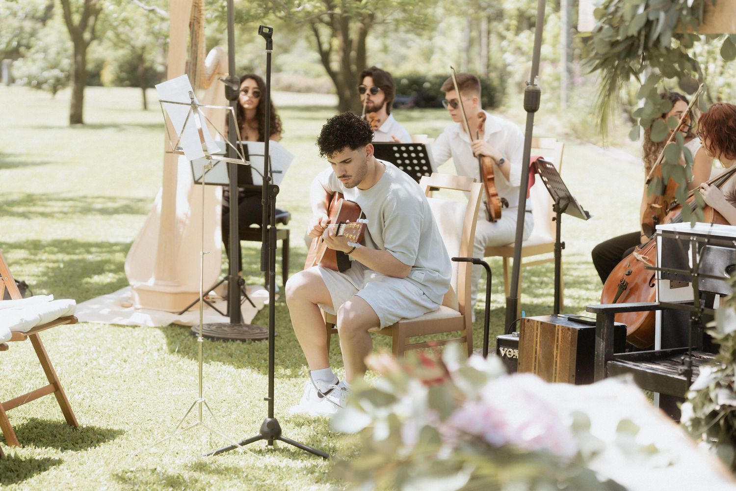 Musicians perform an outdoor concert in a sunny park setting with instruments and music stands.