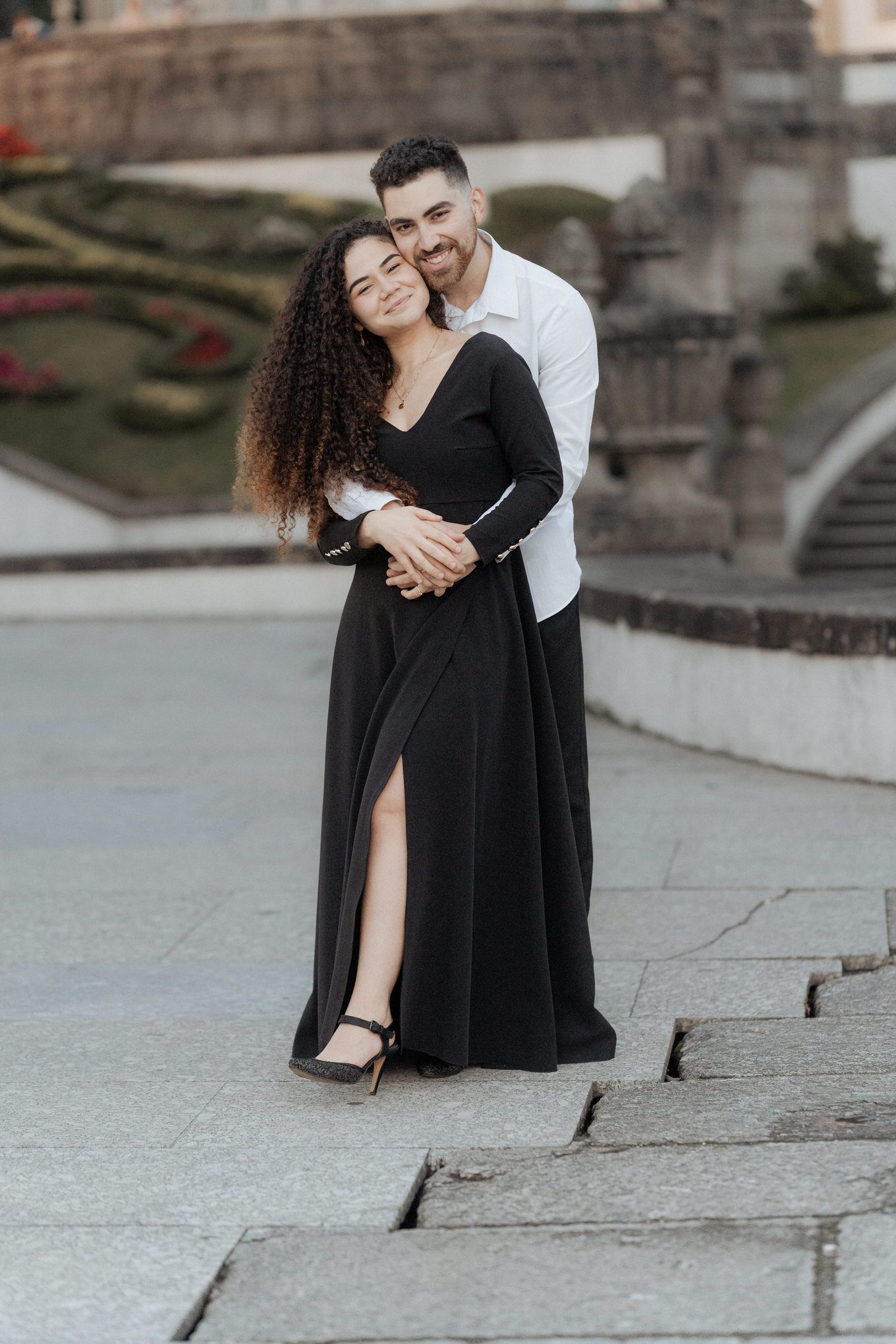 Romantic couple embracing on stone steps, woman in elegant black dress with slit and man in white shirt.