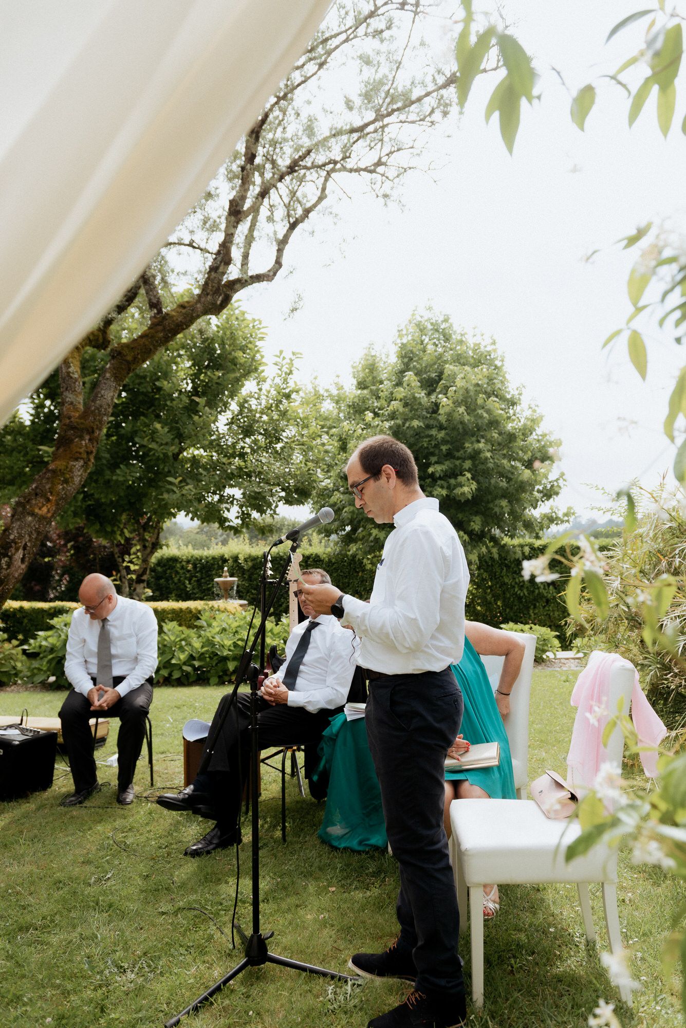 Someone giving a speech at an outdoor wedding ceremony with musicians seated in chairs on a sunny garden lawn.