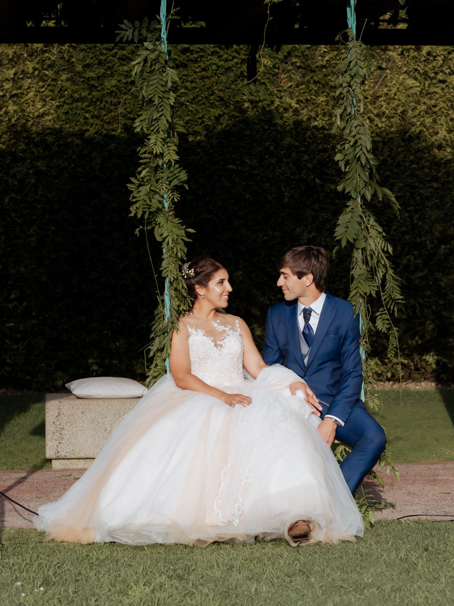 A couple in wedding attire sits on a stone path under willow trees during an outdoor garden photoshoot.