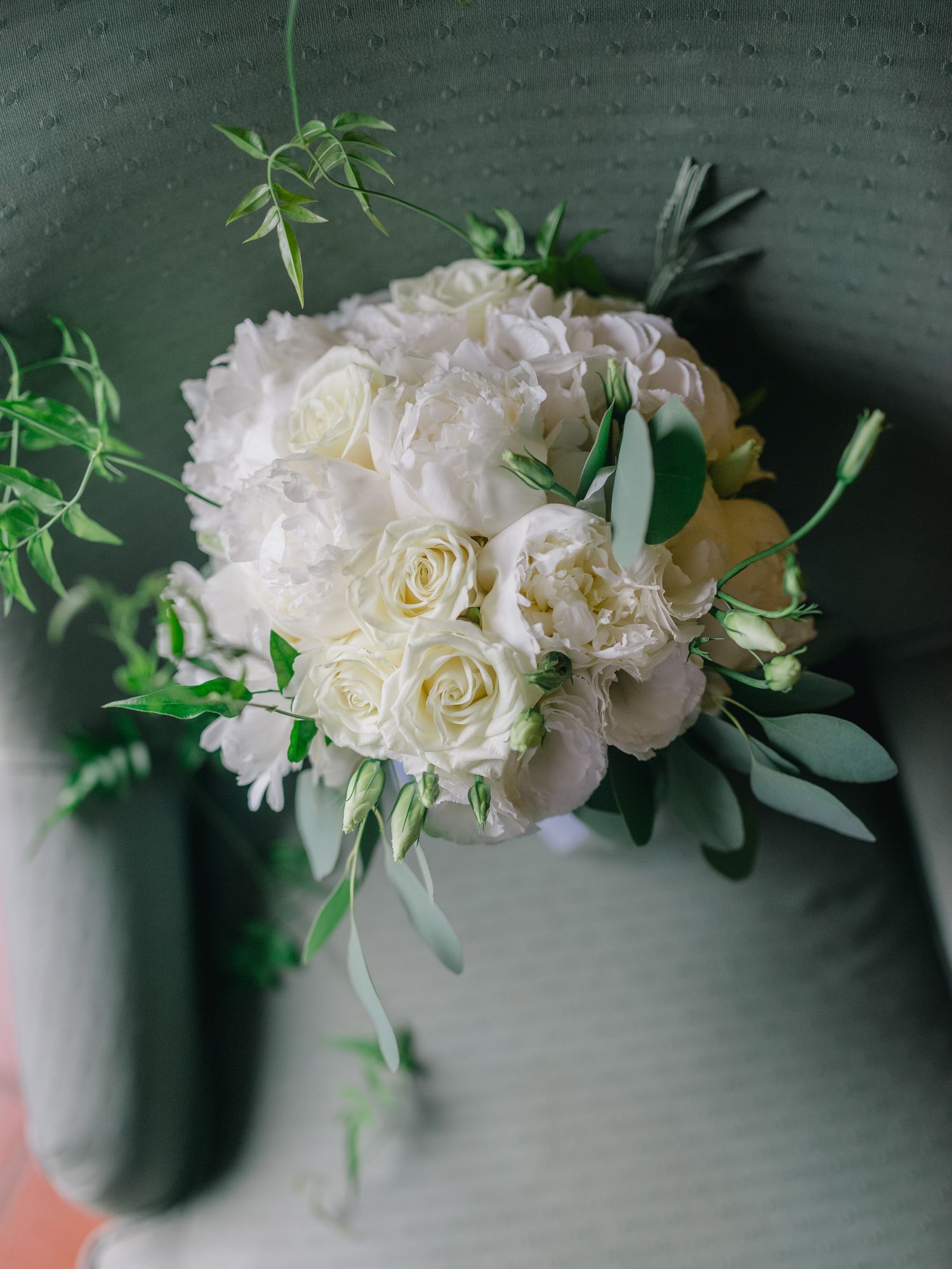 Elegante buquê de rosas brancas e peônias com delicados detalhes verdes dispostos em um fundo escuro texturizado. Detalhes de um casamento.