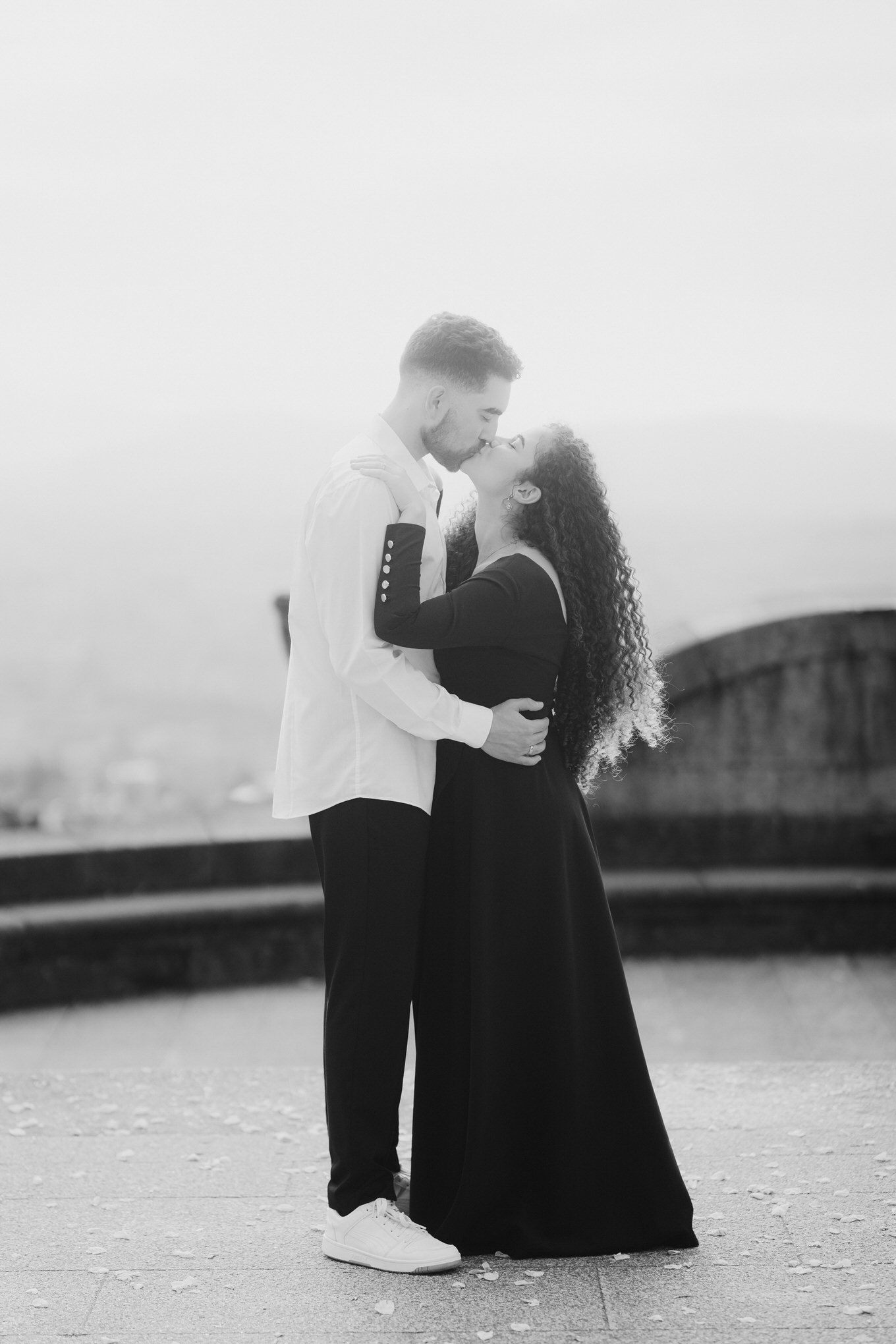 Retrato romântico em preto e branco de um casal se abraçando em um mirante com neblina, ela usando um vestido longo e ele de branco.