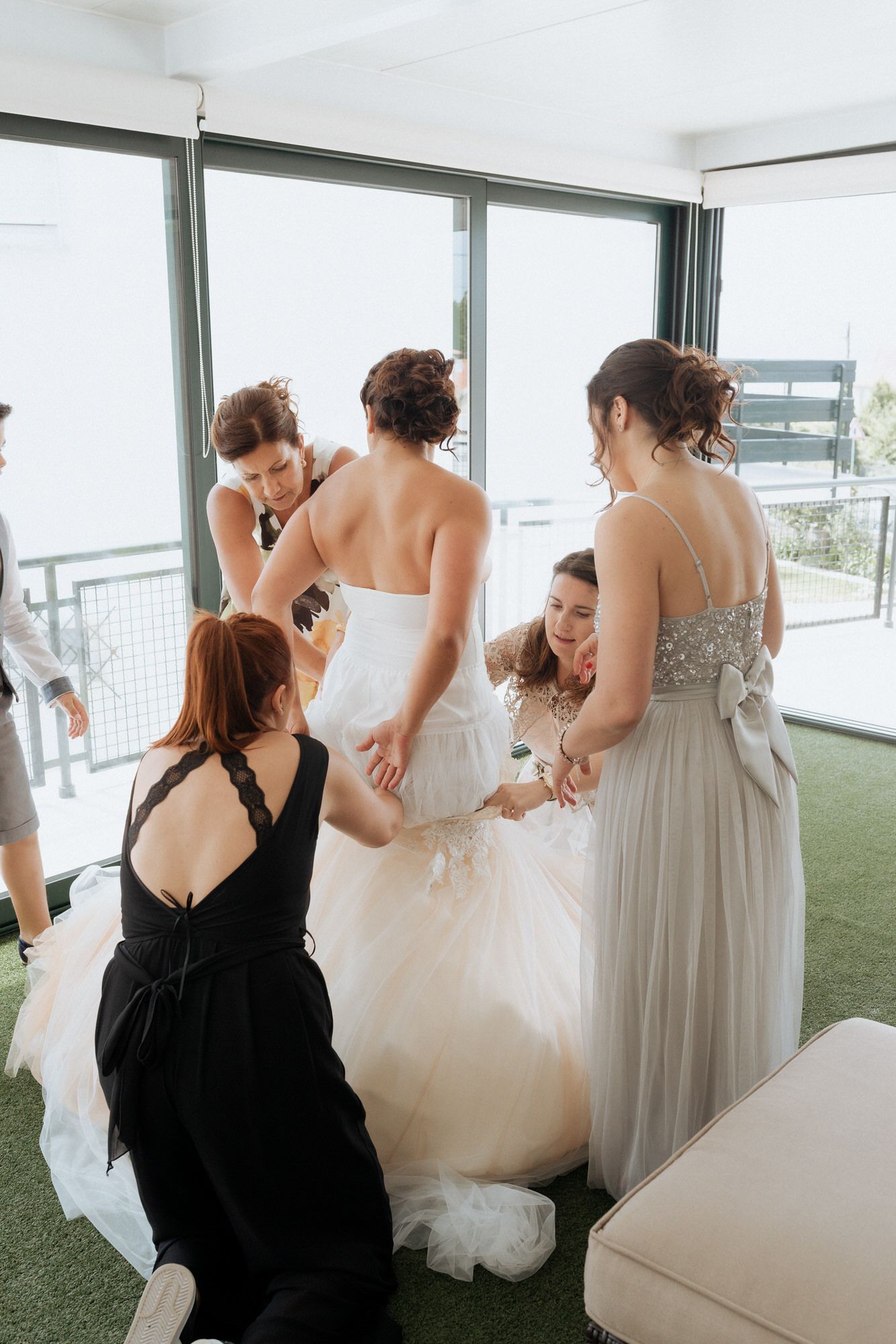 Wedding preparations with bridesmaids helping adjust a flowing white bridal gown in a bright modern room.