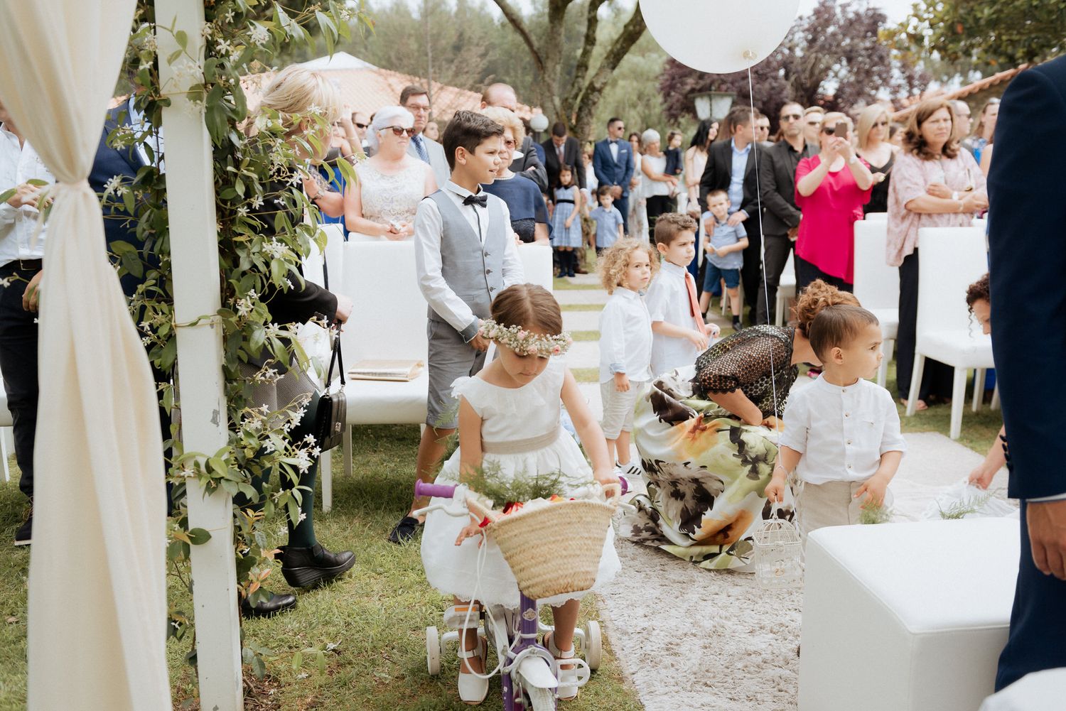 Young flower girls walk down an outdoor wedding aisle path carrying baskets, surrounded by guests at a garden ceremony.