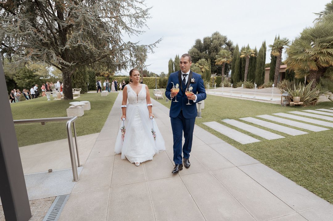 A bride and groom walk hand in hand down a path through a scenic outdoor garden setting on their wedding day.