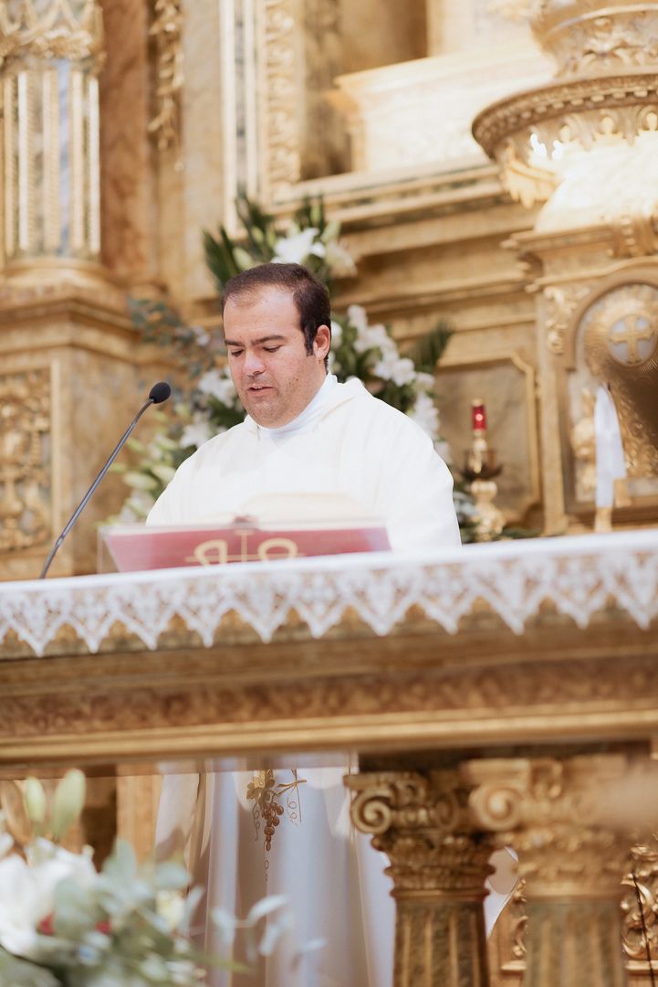 A priest in white robes reads from a book at an ornate golden church altar during a wedding ceremony.