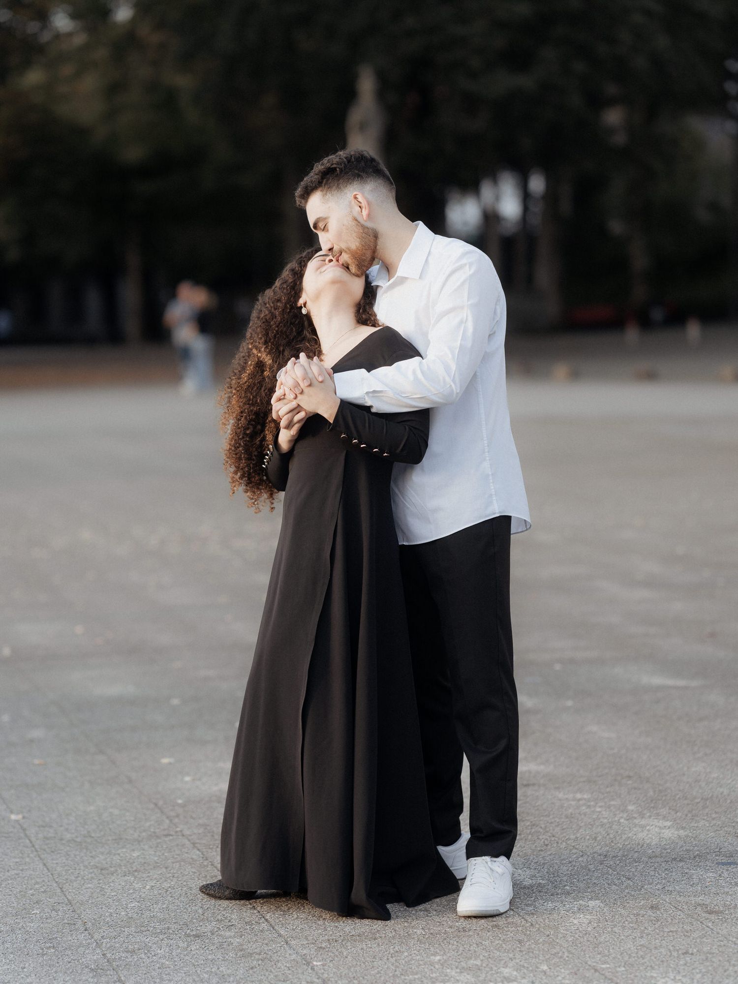 Um casal romântico se abraçando em trajes formais, com a mulher usando um vestido longo escuro e o homem com uma camisa branca ao ar livre.