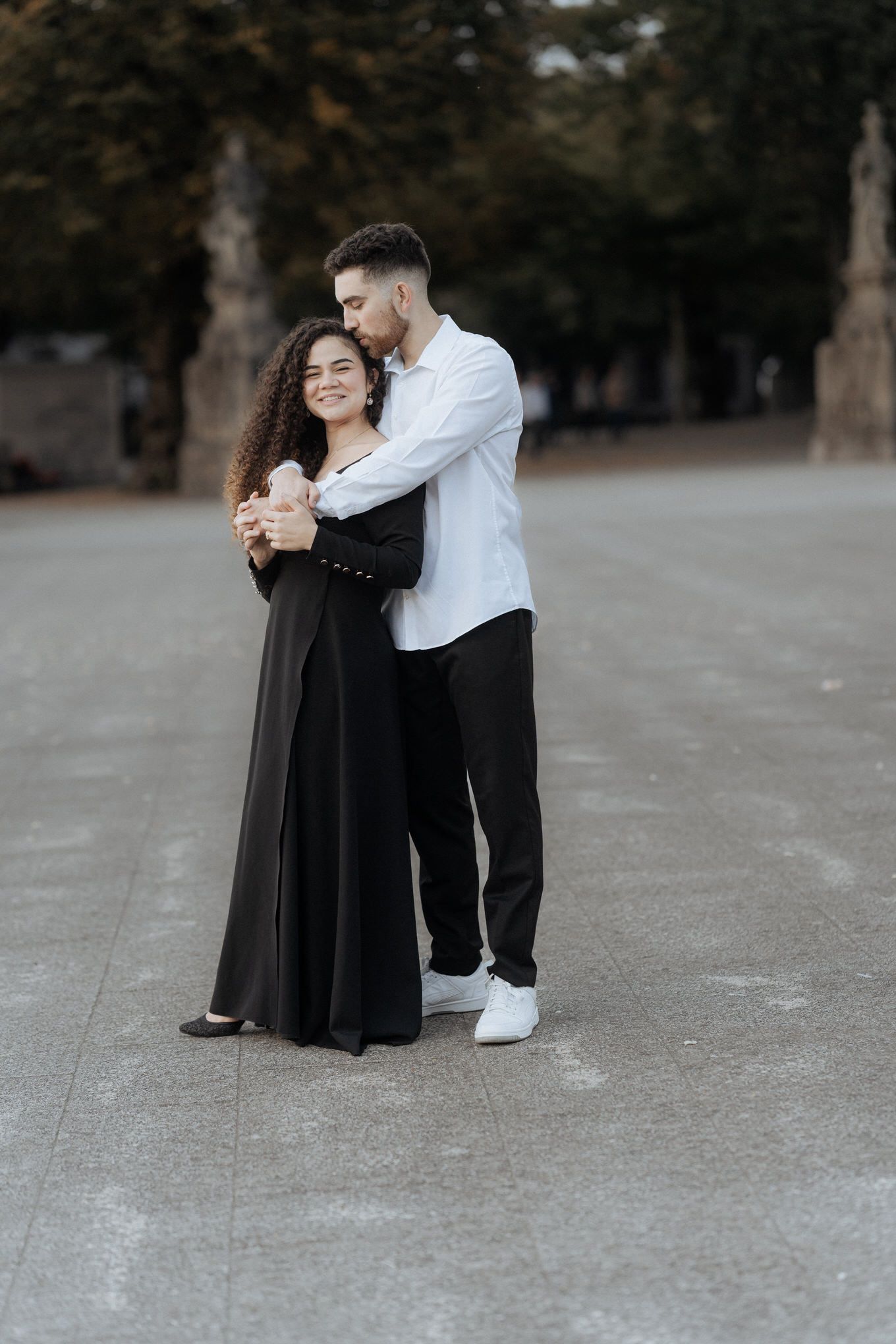 Um casal romântico se abraça ao ar livre, um usando um vestido preto e o outro uma camisa branca e calças escuras.