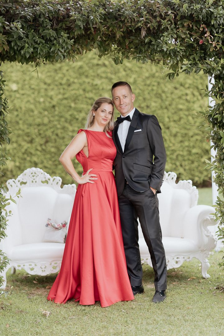 Couple poses outdoors in formal attire with white vintage furniture and greenery in a garden setting.